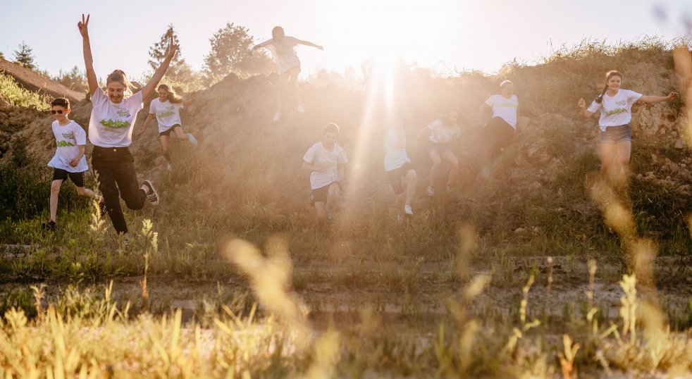 Grupa młodych ludzi zbiega ze skarpy, jednocześnie radośnie podskakując. Pomiędzy nimi przebijaą się promienie słońca. Mają na sobie koszulki Programu 
